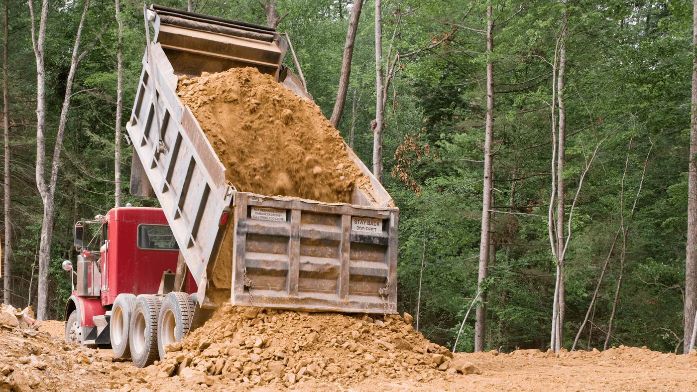 Tandem dump truck dumping a load of fill dirt on a wooded site